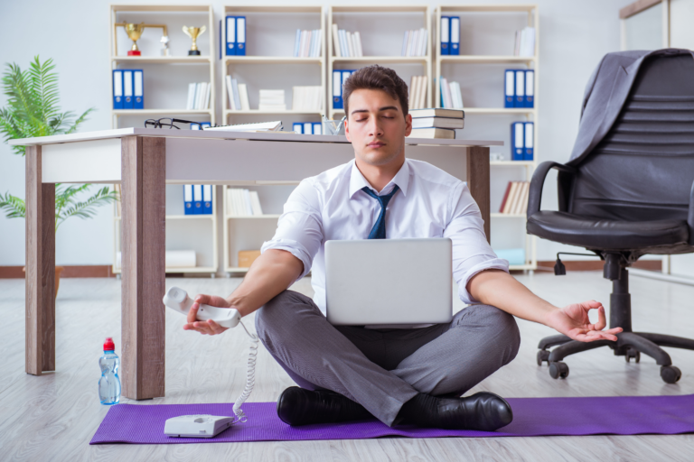 Man op kantoor zit in meditatiehouding. Hij heeft een laptop in zijn schoot en telefoon in zijn hand.